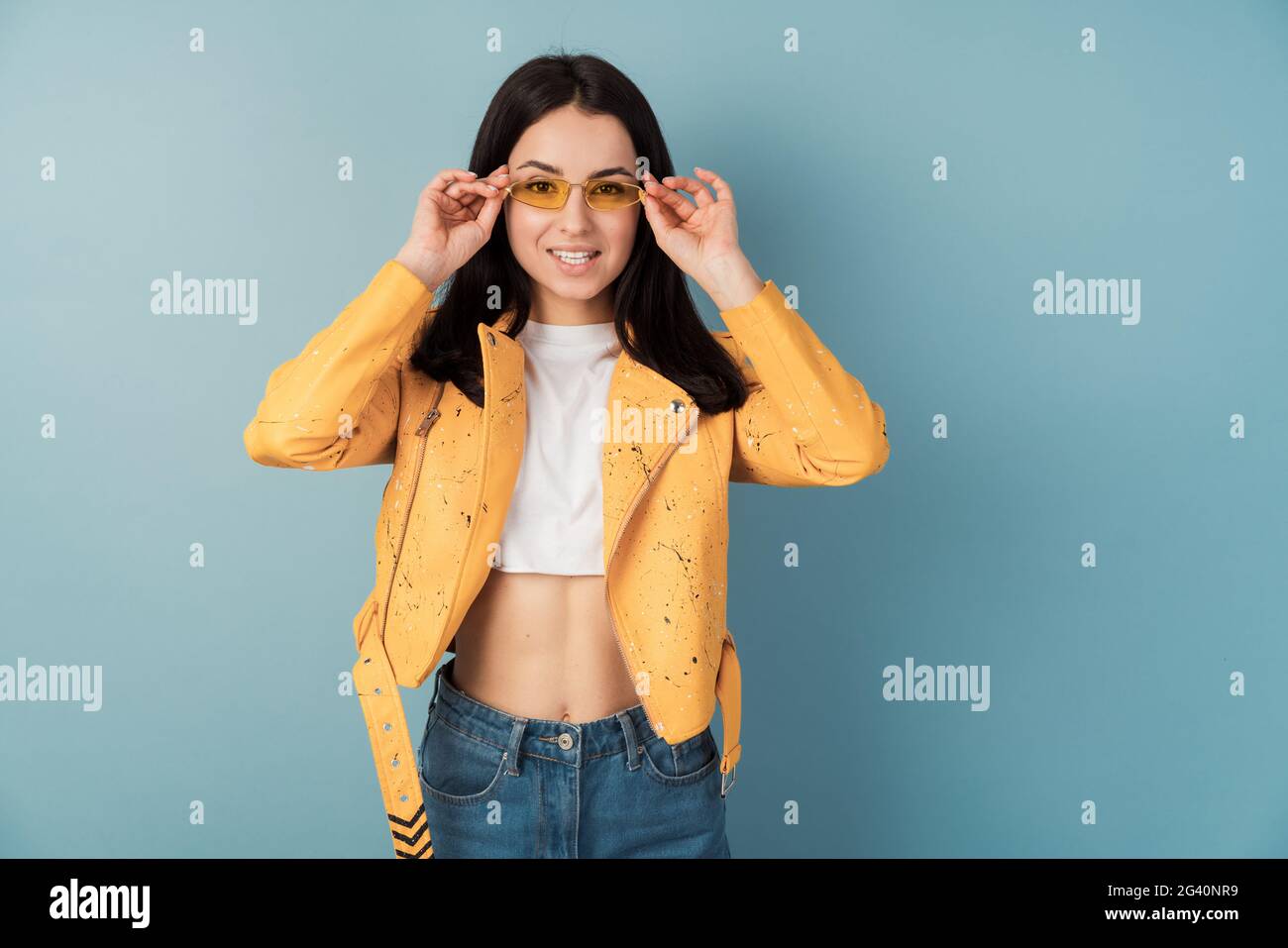 Positive girl adjusts sunglasses, she wears an orange casual jacket and blue jeans. Pretty lady on blank wall background, copy space, place for text. Stock Photo