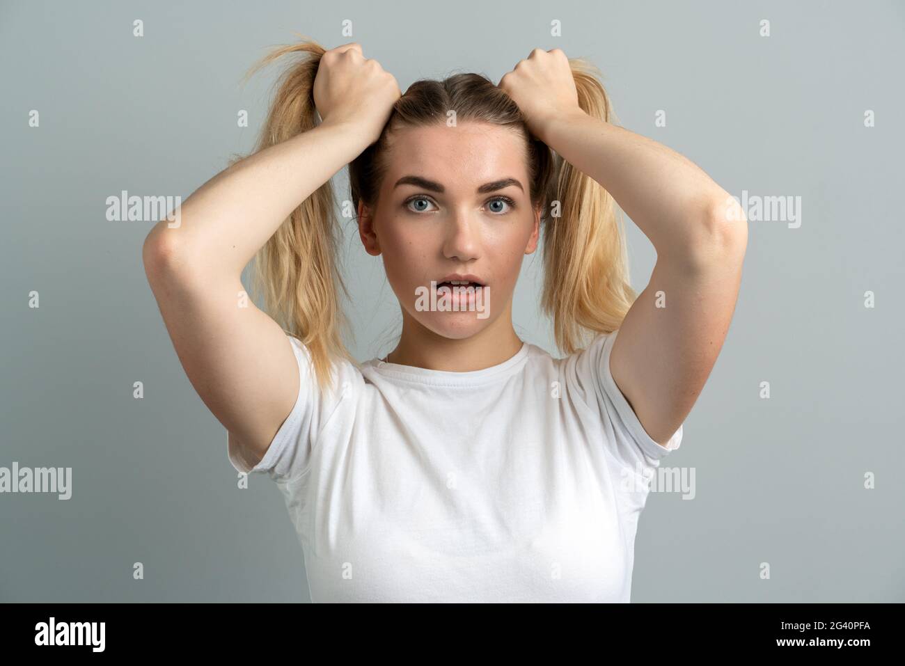 Girl with a shocking look on a gray background. Beautiful, young girl holding two ponytails from her hair. Stock Photo