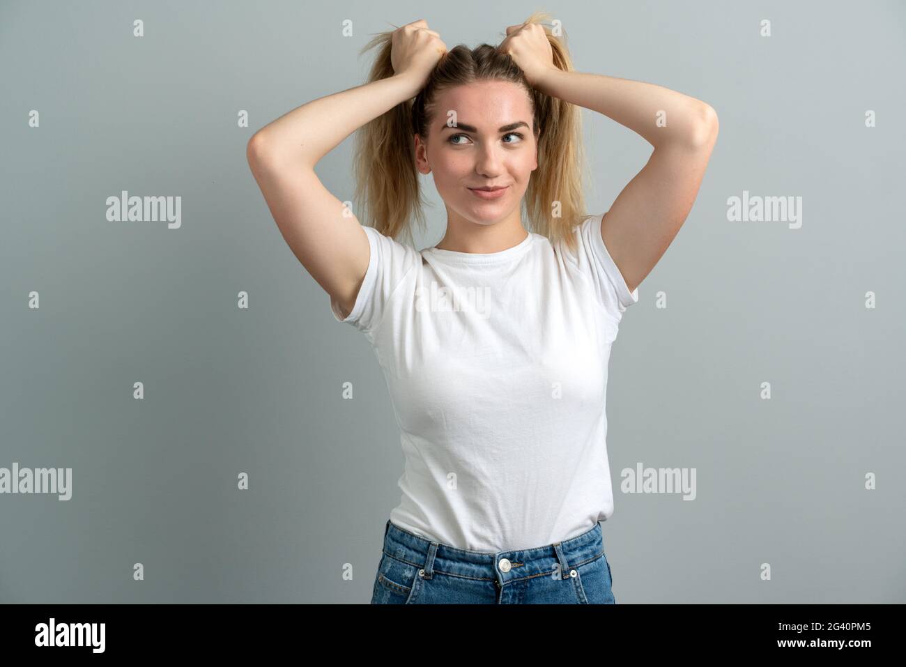Funny playful young woman hold her blonde hair into two ponytails. Posing on camera on gray background. Stock Photo