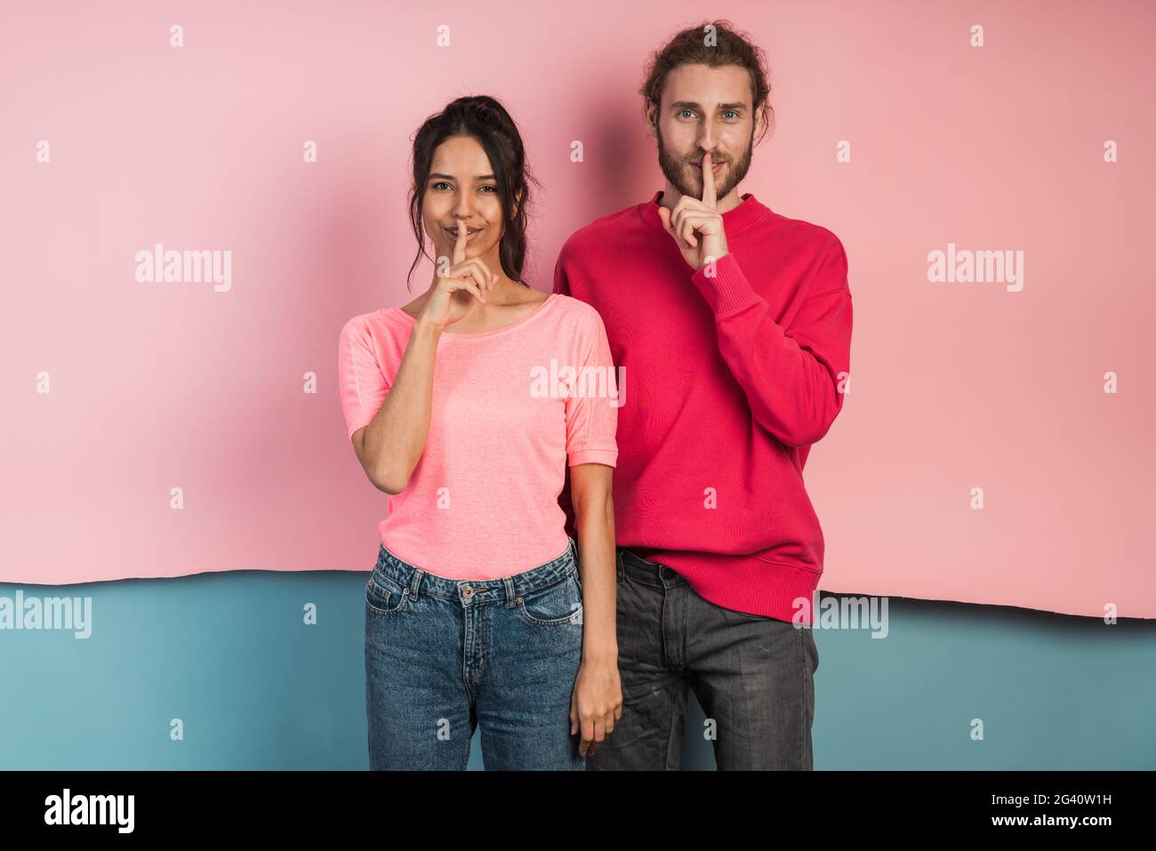 Attractive young couple making a gesture to be quiet. They touch the index fingers of their mouths on a pink and blue background. Stock Photo