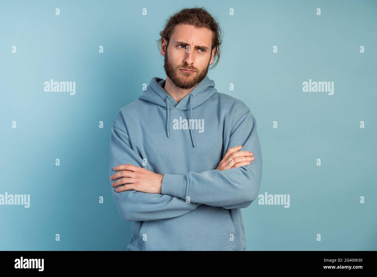 Photo of an upset man in a hooded sweater, he crossed his arms over his chest. Man posing on a blue background. Stock Photo