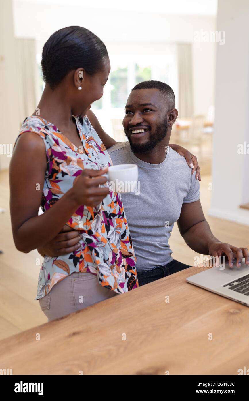 Happy african american couple embracing in kitchen, man using laptop ...