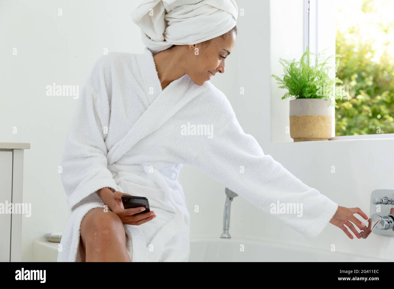 Mixed race woman wearing bathrobe sitting on bathtub edge and using smartphone in bathroom Stock Photo
