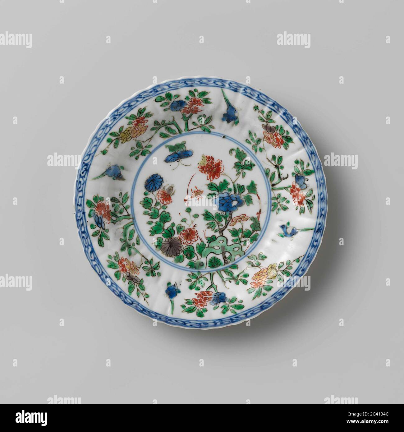 Fluted Saucer With Rocks, Flower Sprays and Birds. Dish of porcelain with ribbed wall, partly covered with café-au-lait, painted in underglaze blue and on the glaze blue, red, green, yellow, eggplant, black and gold. On the flat a butterfly and flowering plants (peony, chrysanthemum) at a rock; Four flour branches on the wall interspersed with a bird; the edge with curlwork; The rear is covered with a café-au-lait-colored glaze with three flower branches. Marked on the bottom with a mushroom (Lingzhi) [?] In a double circle. Famille Verte. Stock Photo