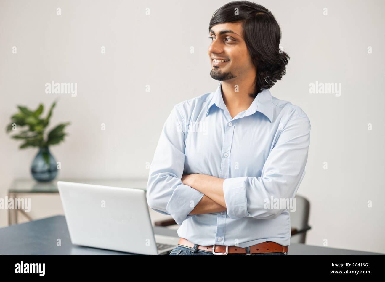 High skilled indian man, ceo, male employee in formal wear stands with folded arms in bright office space, looks away and planning new aims and goal. Aspiration and success concept Stock Photo