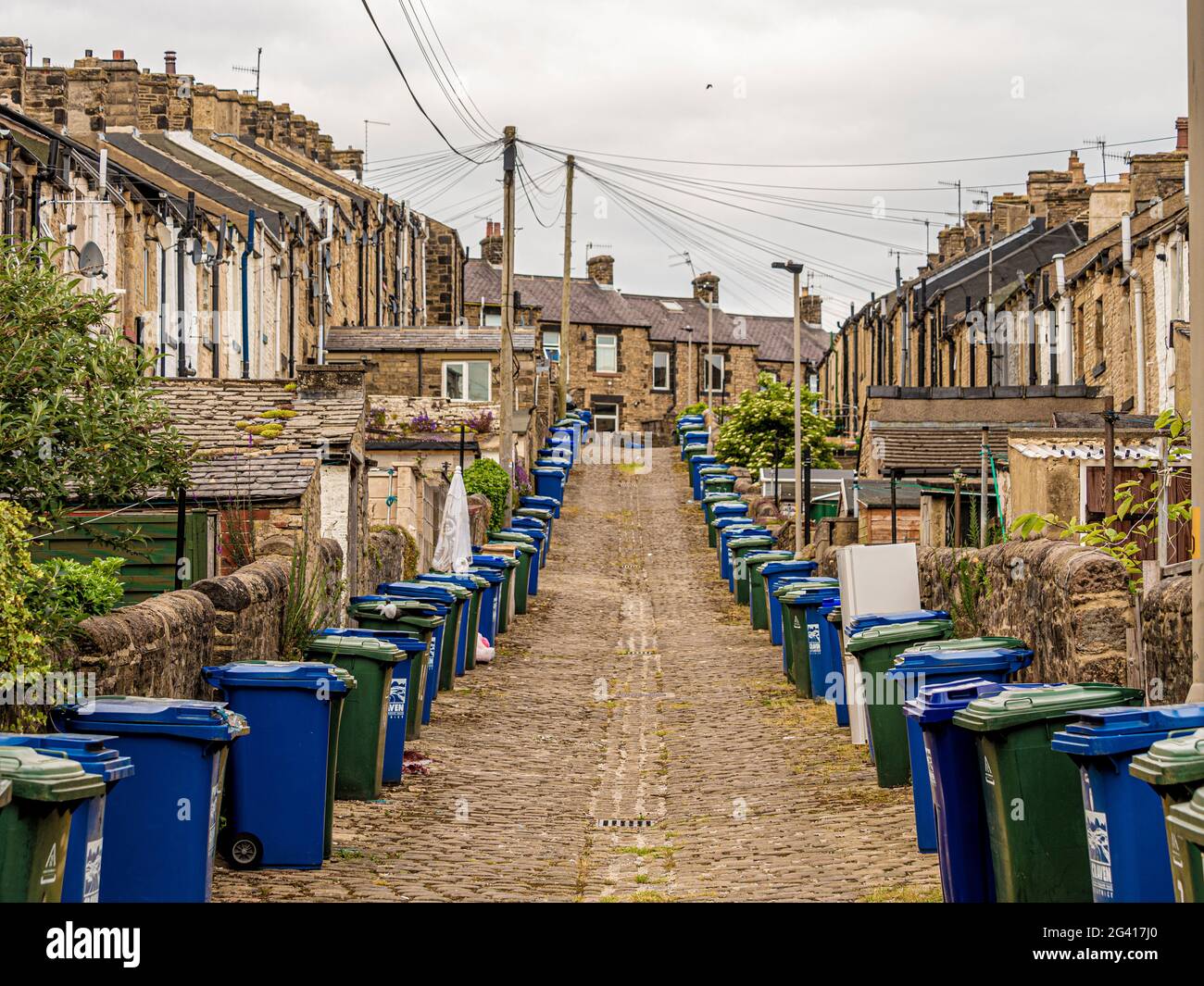 Sloping cobbled back alley at rear of Victorian terraced houses in Skipton, UK, with lines of wheelie bins. Stock Photo