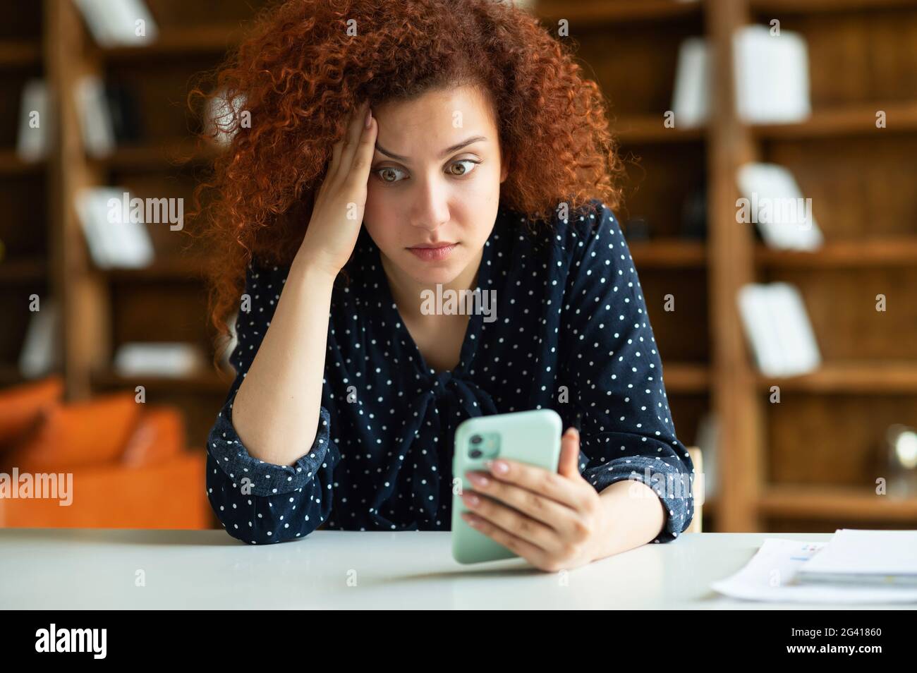 Worried red-haired young female freelancer or student with curly hair sitting at the desk, holding smartphone with puzzled face expression, received unexpected bad shocked news Stock Photo