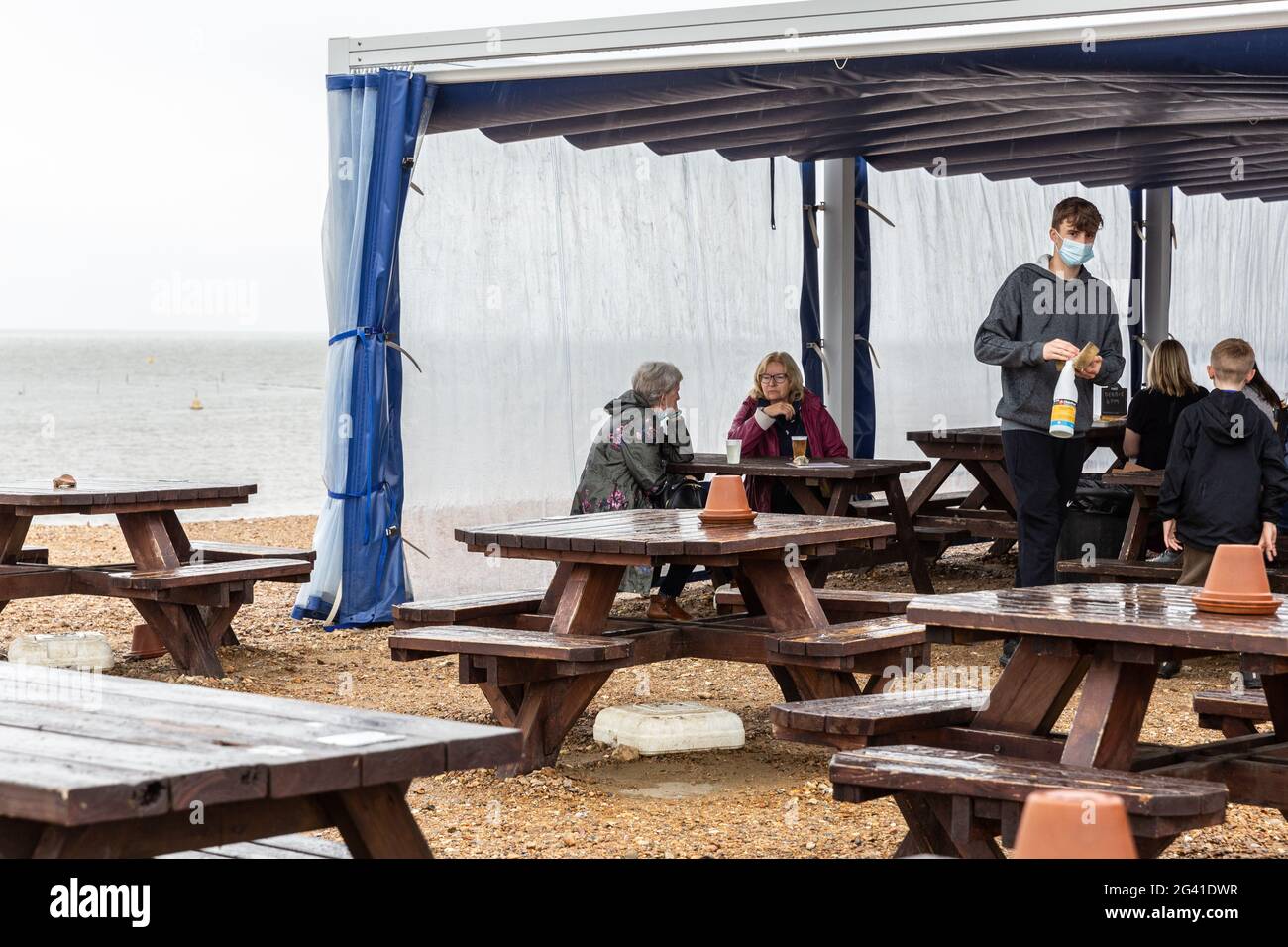 Whitstable, United Kingdom, June 18, 2021. People  sit in outdoor restaurant at the beach  in Whitstable, a coastal touristic town in south-eastern England as vary poor rainy weather disturb holidaymakers as the holiday season begins. One of the strictest Coronavirus lockdowns in the world  is partially lifted as the United Kingdom managed to vaccinate a large part of the population and the number of Covid cases is low. People are able to socialize in limited numbers and are able to book holidays. Stock Photo