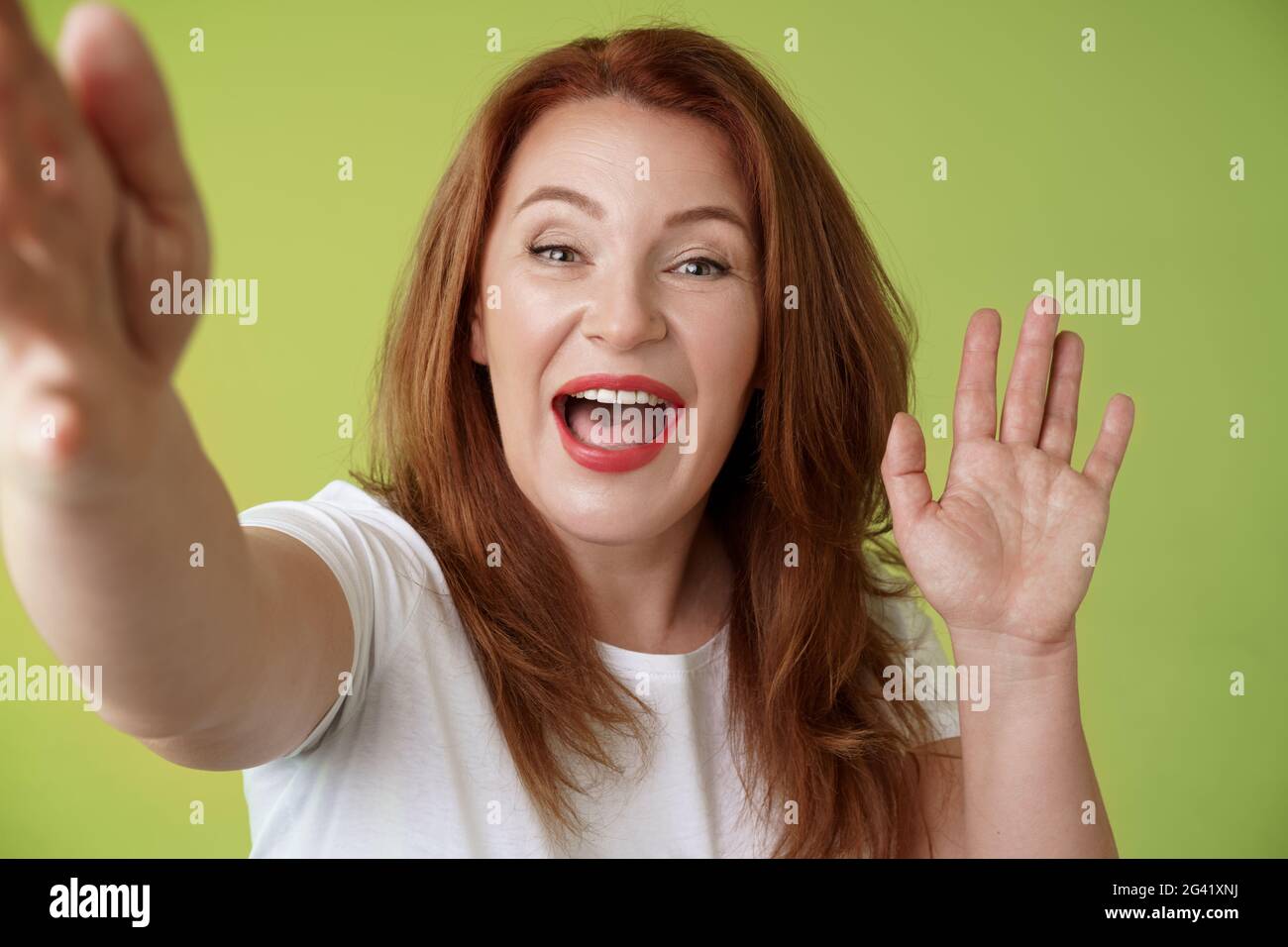 Friendly cheerful redhead middle-aged woman extend arm hold camera taking selfie waving palm hi hello greeting smiling broadly w Stock Photo