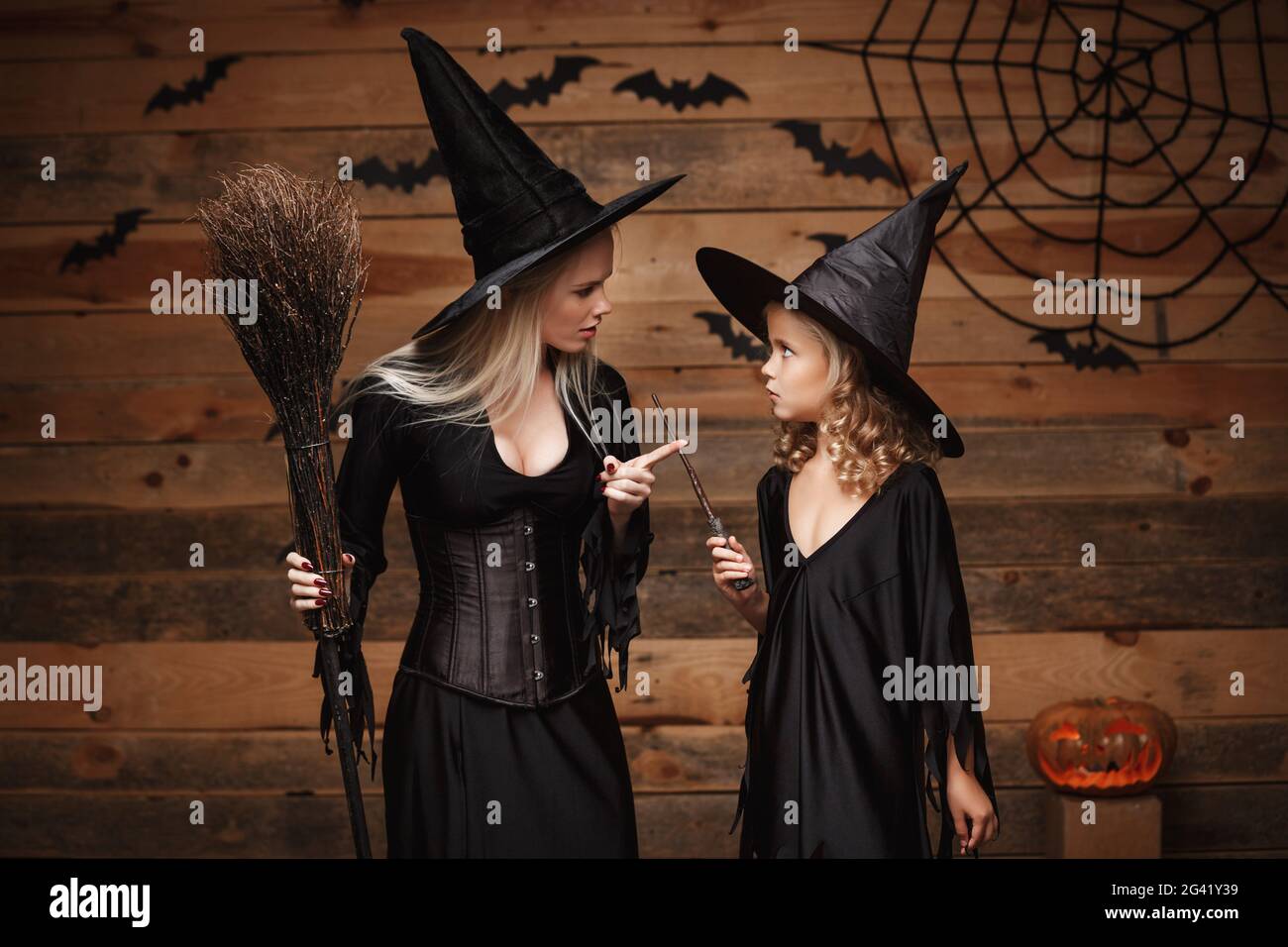 Halloween Concept - stressful witch mother teaching her daughter in witch costumes celebrating Halloween with curved pumpkins ov Stock Photo