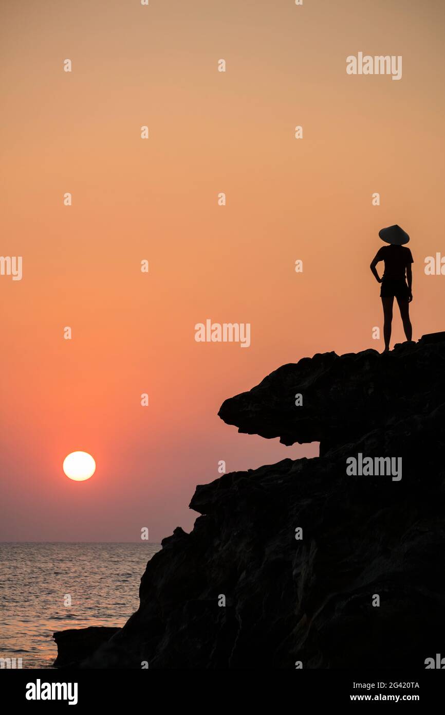 Silhouette of young woman wearing conical hat and looking out to sea from rock ledge next to Dinh Cao Shrine at sunset, Duong Dong, Phu Quoc Island, K Stock Photo