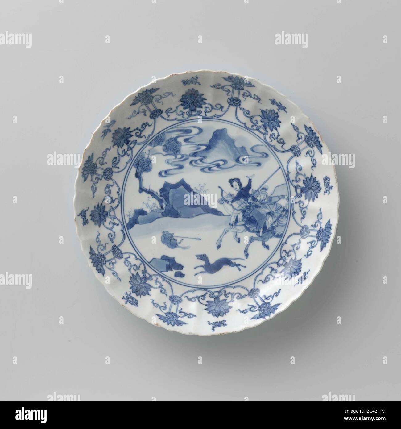 Saucer-dish with hunting scene, Lotus Scrolls and auspicious Symbols. Scale of porcelain with round, ribbed wall and scalloped edge, painted in underglaze blue. On the flat a hunting scene with two armed Saucer-dish with hunting scene, Lotus Scrolls and auspicious Symbols. Scale of porcelain with round, ribbed wall and scalloped edge, painted in underglaze blue. On the flat a hunting scene with two armed