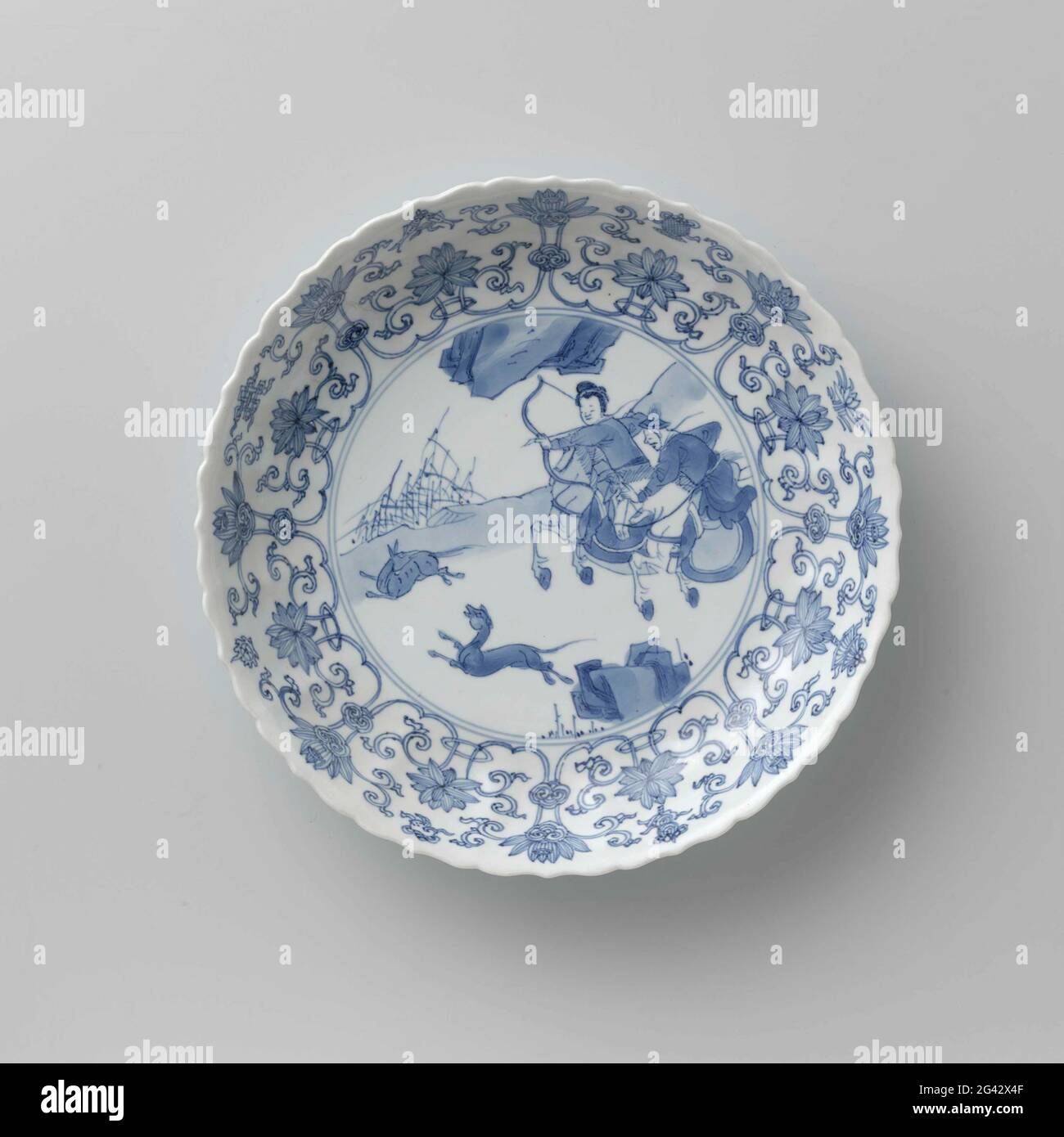 Saucer-dish with hunting scene, Lotus Scrolls and auspicious Symbols. Scale of porcelain with round, ribbed wall and scalloped edge, painted in underglaze blue. On the flat a hunting scene with two armed Saucer-dish with hunting scene, Lotus Scrolls and auspicious Symbols. Scale of porcelain with round, ribbed wall and scalloped edge, painted in underglaze blue. On the flat a hunting scene with two armed