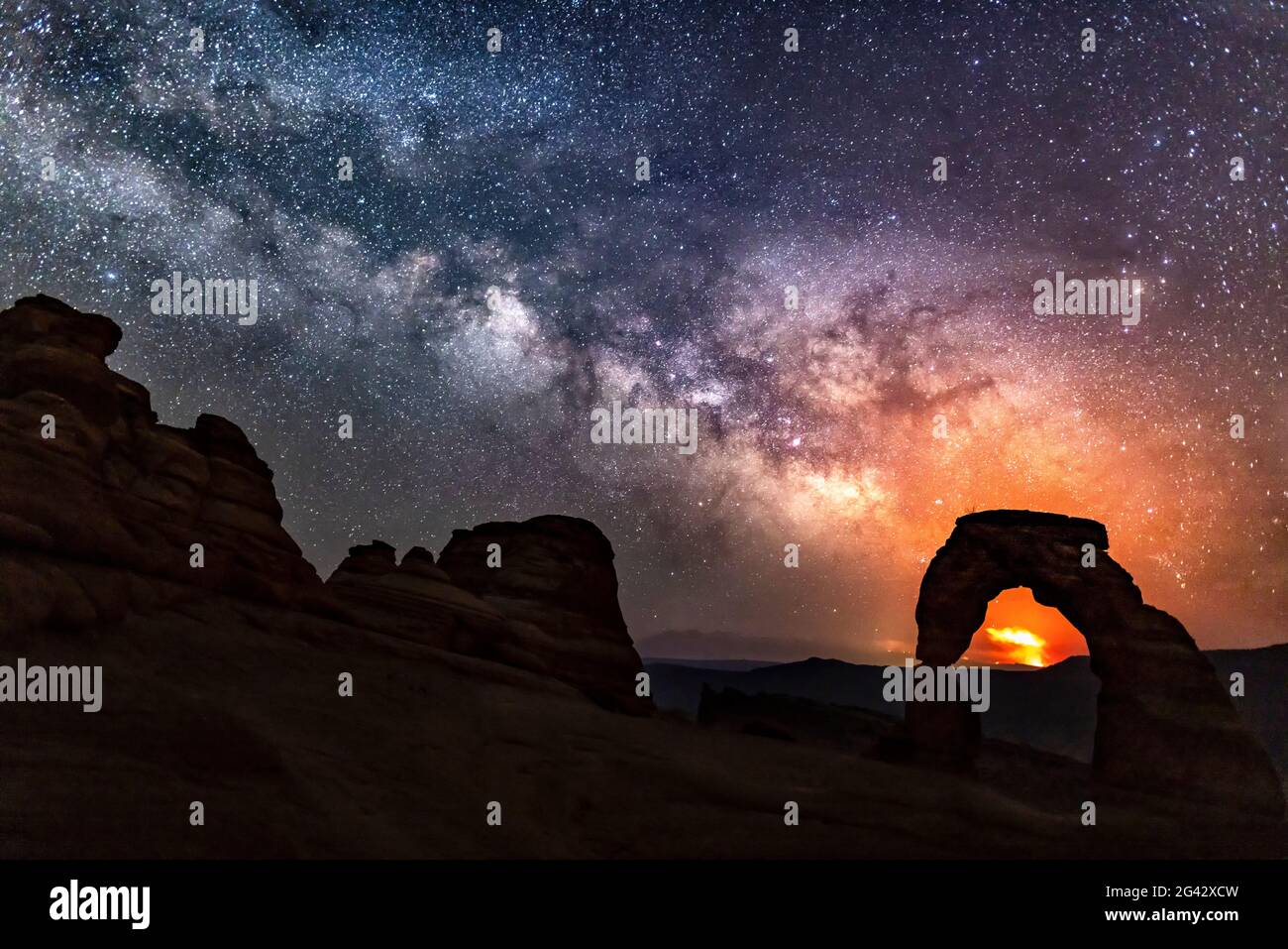 The Pack Creek Fire in the La Sal Mountains lights up the night sky in the distance, with the Milky Way over Delicate Arch in Arches National Park, Ut Stock Photo