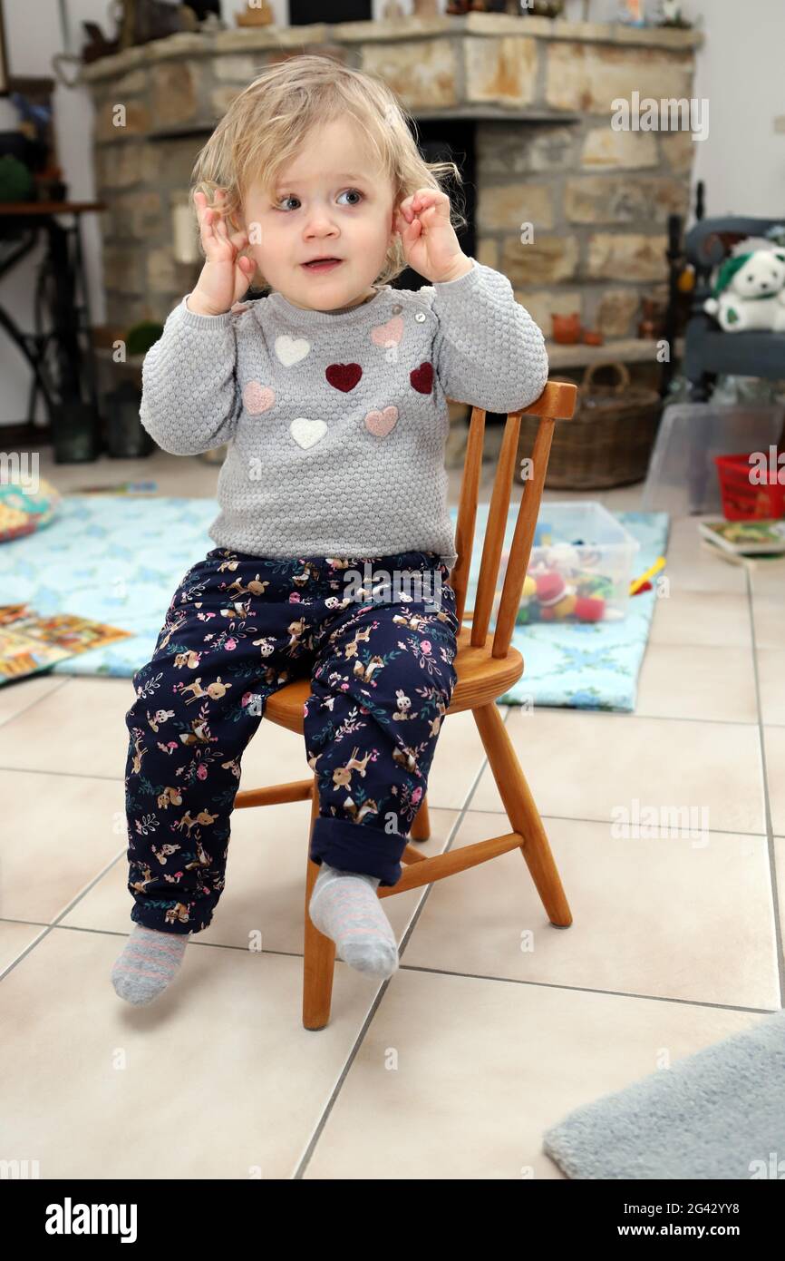 Little girl sitting on a wooden child chair in living room and covers her ears Stock Photo
