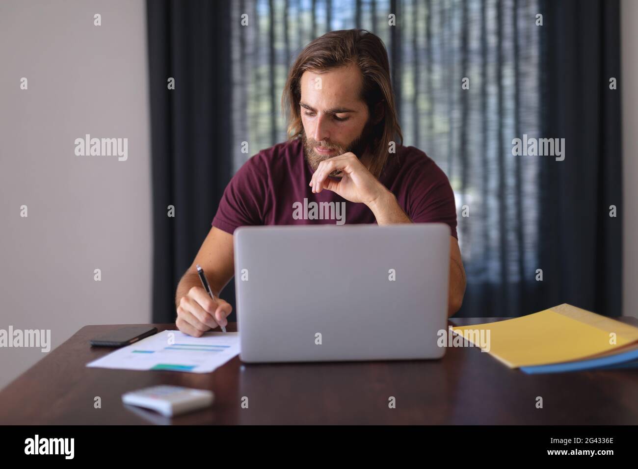 Caucasian man sitting at table working from home, making notes and using laptop Stock Photo