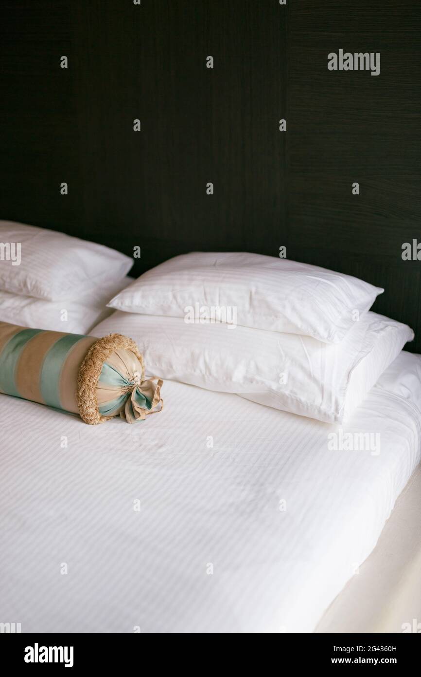 A bed with a white blanket and pillows of different sizes and an elongated cushion in a room with dark walls. Stock Photo