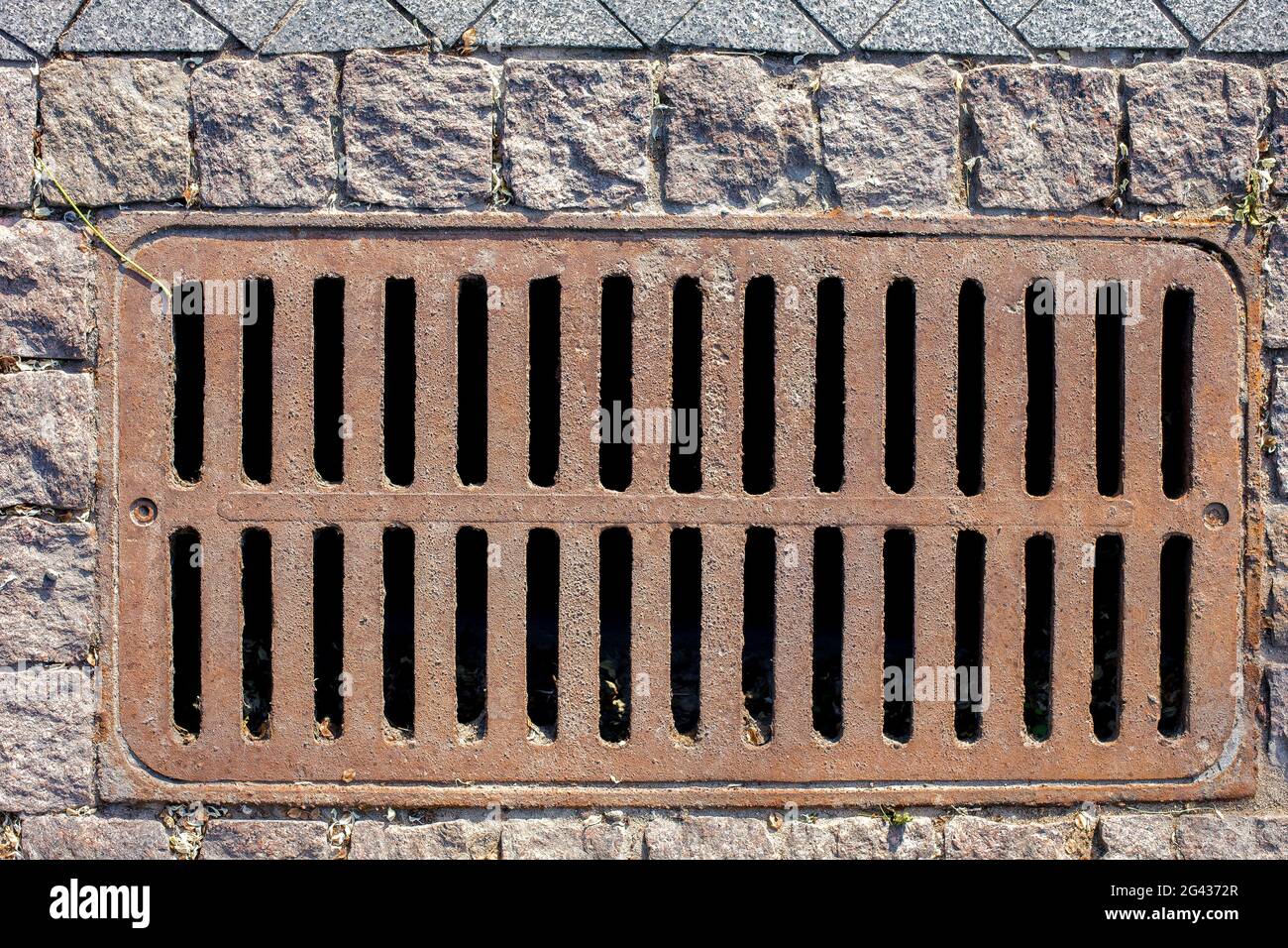 rusty manhole rectangular grating of the drainage system of the pedestrian sidewalk from rough cut stone tiles top view. Stock Photo