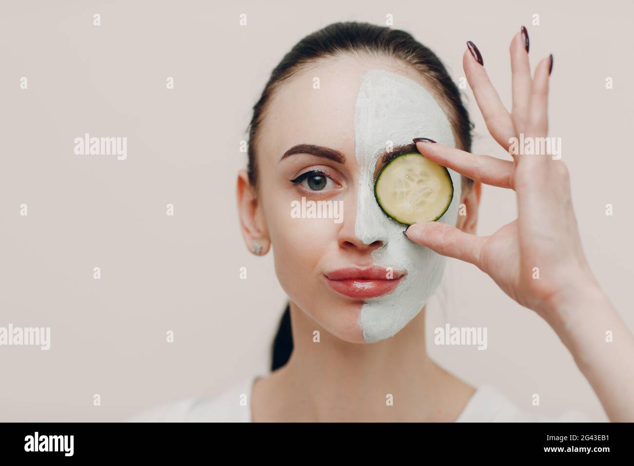 Young woman applying mask of clay on face with cucumber on eyes in beauty spa Stock Photo
