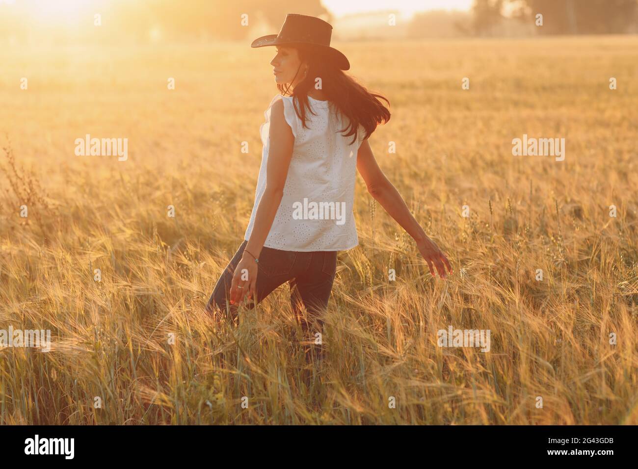 Woman farmer in cowboy hat walking with hands on ears at agricultural field on sunset Stock Photo