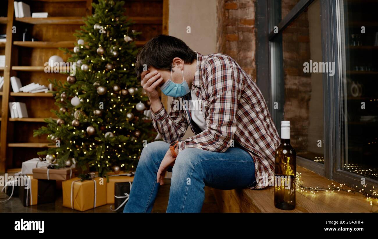 Lonely man wearing Covid 19 face mask very upset and drunk, sitting by window and Christmas tree and drinking alcohol from bottl Stock Photo