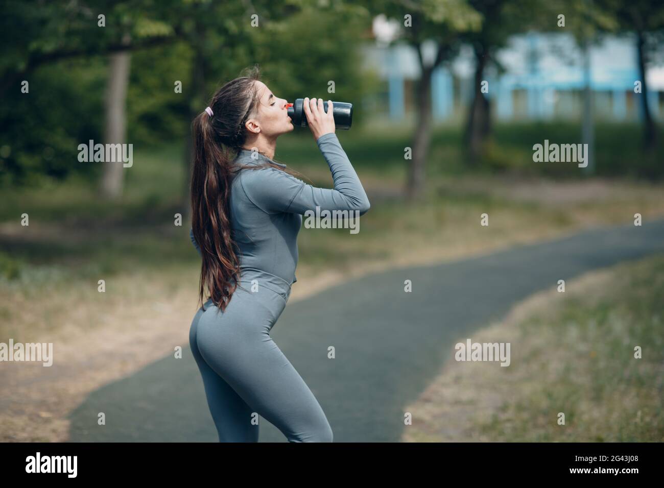 Tired woman jogger in gray tracksuit drinking bottled water after jogging in park Stock Photo