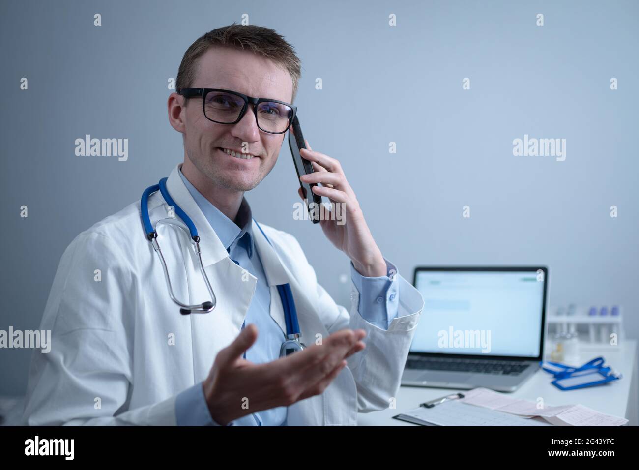 Smiling doctor having phone call at desk. Physicianr talking with his patient in office clinic sitting at table with laptop. Ger Stock Photo