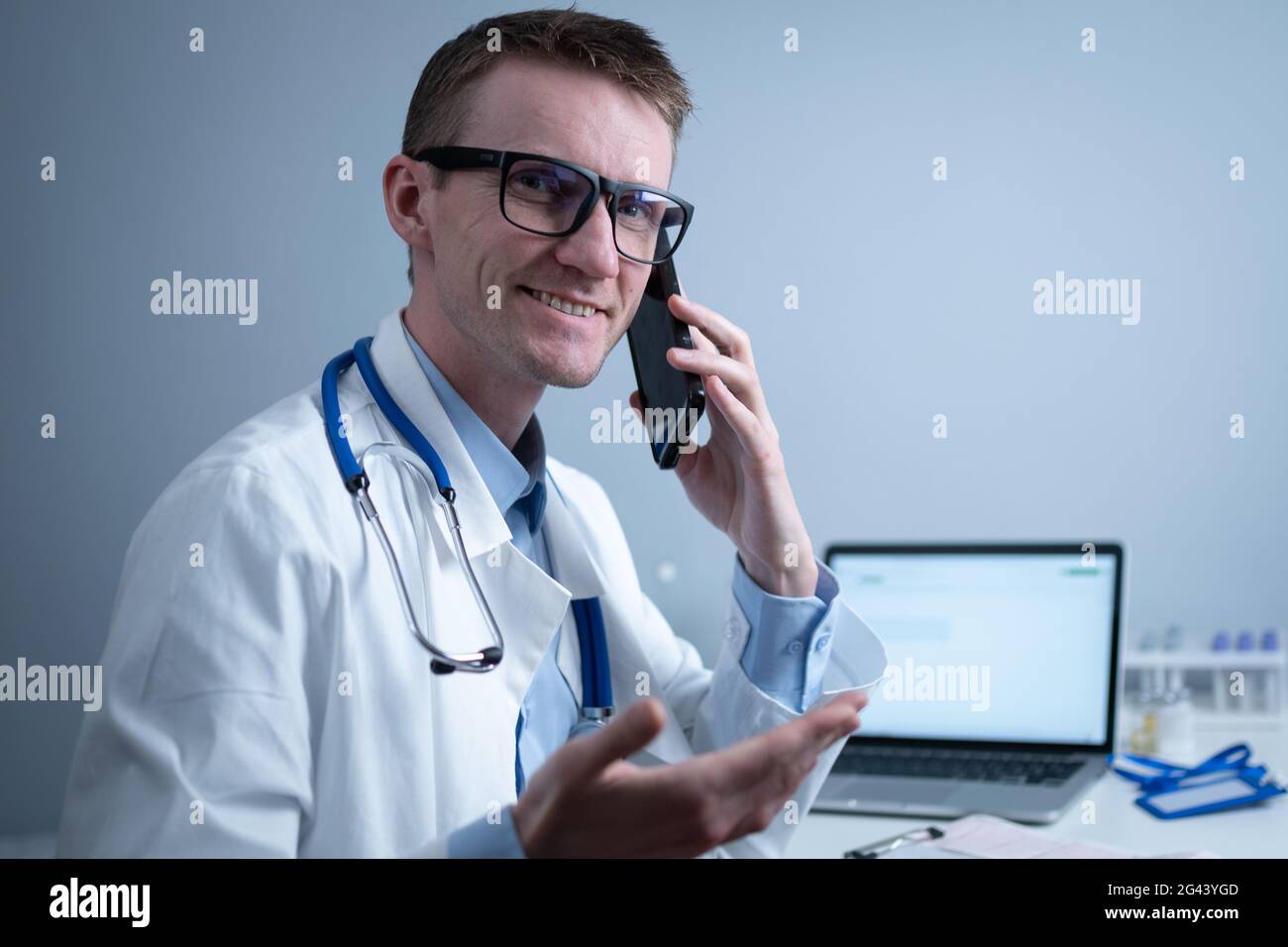 European doctor in white medical coat and glasses consults patient on cell phone in clinic office, sitting at table with laptop. Stock Photo