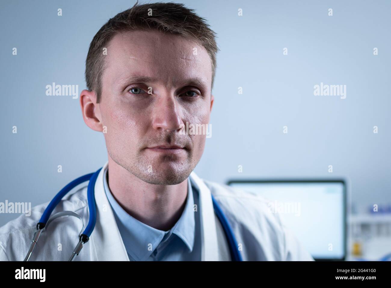 Tired young doctor looking at camera in hospital office on background of desktop. Topic is overburdened medical system at corona Stock Photo