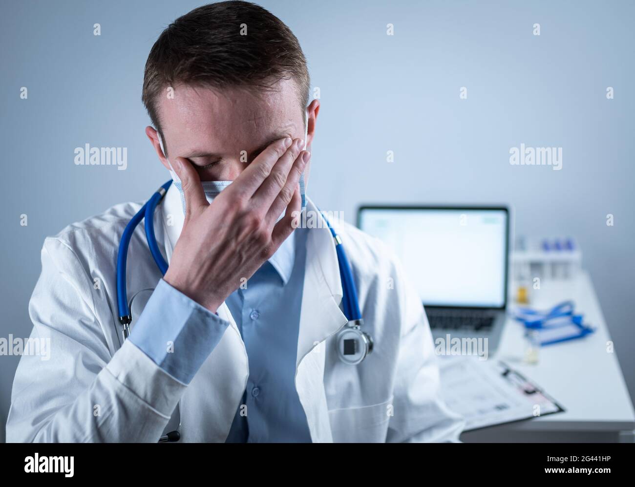 Exhausted young doctor in lab coat takes off mask and glasses, rubs eyes, has headache from fatigue and overexertion from workin Stock Photo