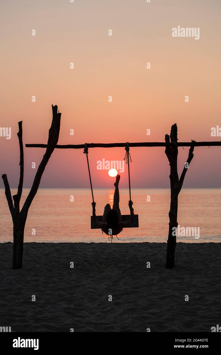Silhouette of young woman on swing at Ong Lang Beach at sunset pretending to be kicking the sun like football, Ong Lang, Phu Quoc Island, Kien Giang, Stock Photo