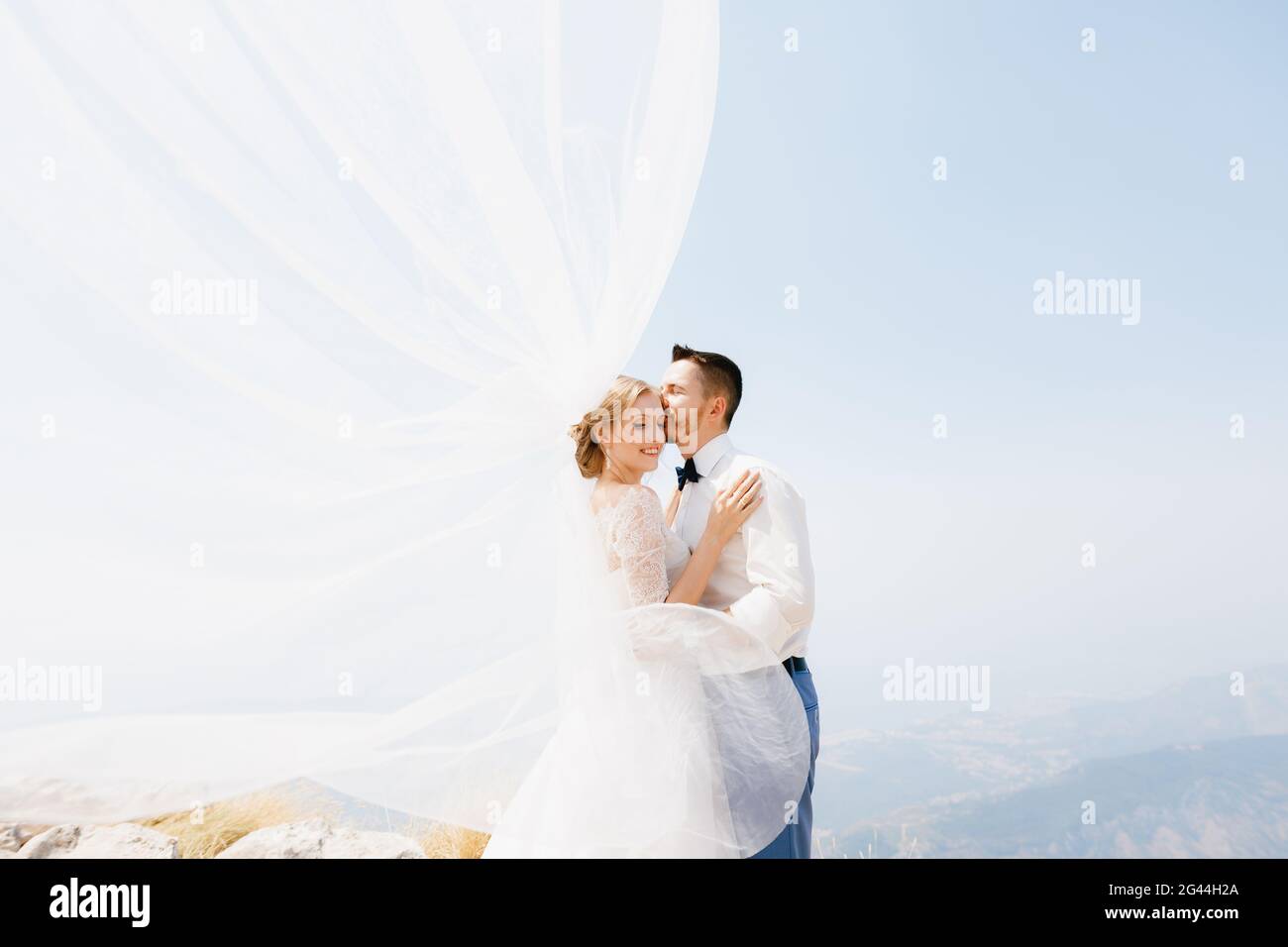 The bride and groom are embracing on the mountain, the groom kisses the bride, the wind waves the veil Stock Photo