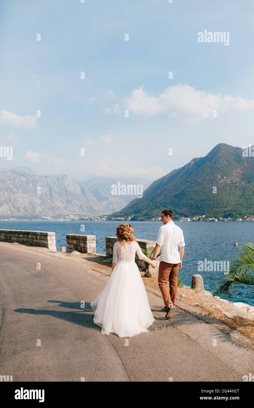 The bride and groom walk hand in hand along the road along the coast in the Bay of Kotor near Perast, back view Stock Photo