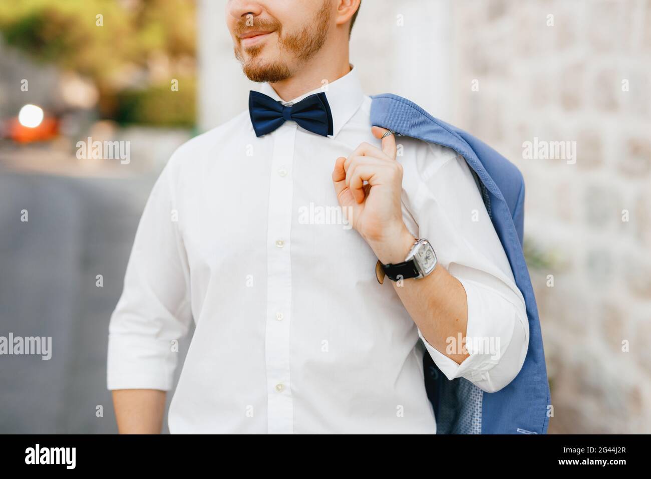 A man stands in front of a white brick house in the old town of Perast and holds a jacket over his shoulder Stock Photo