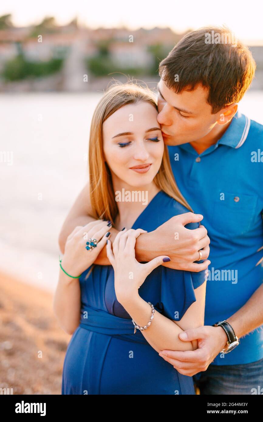 Man gently kisses smiling pregnant woman in a blue dress hugging her shoulders Stock Photo