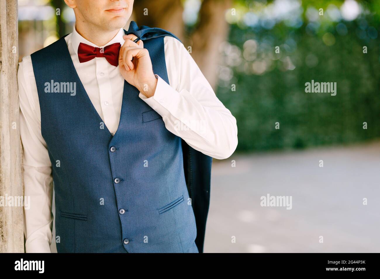 Groom holds his jacket on his shoulder, leaning against a pillar in the garden Stock Photo