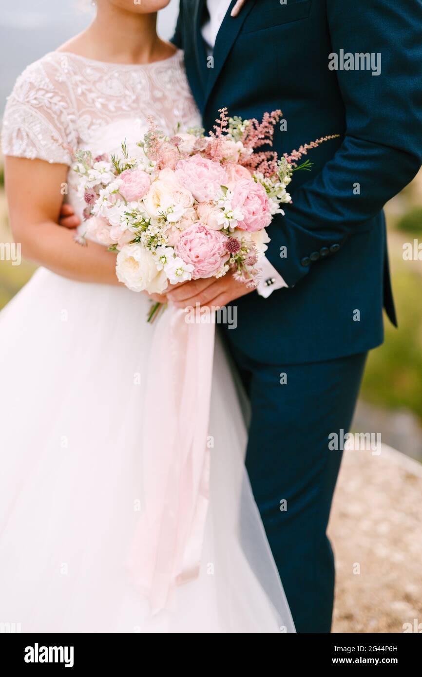 The bride and groom stand hugging and hold the bride's bouquet with delicate pink roses, peonies and astilbe, close-up Stock Photo