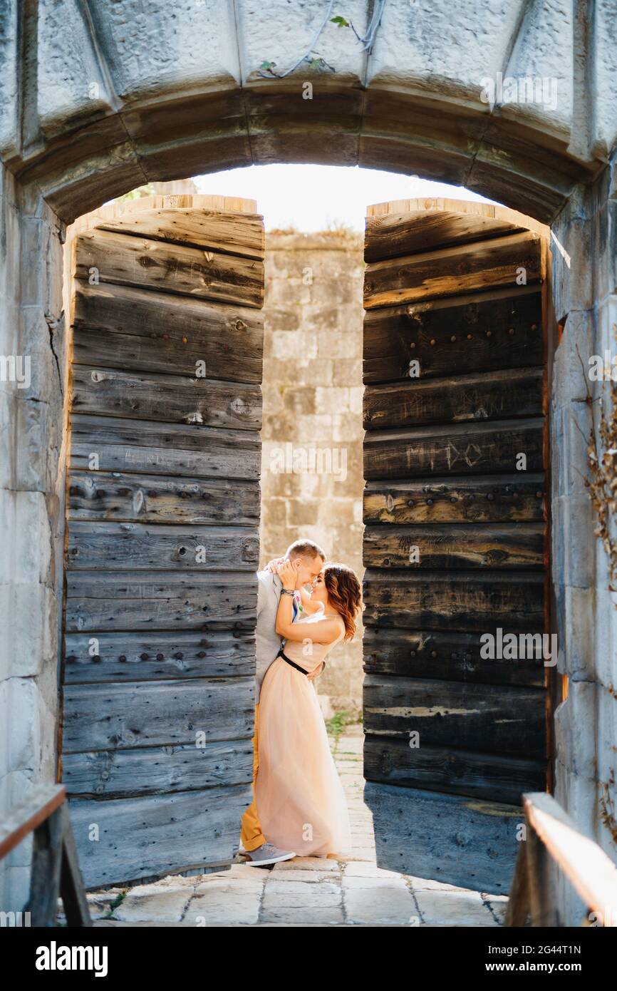 Bride hugs groom while standing behind the open wooden gate of the castle Stock Photo