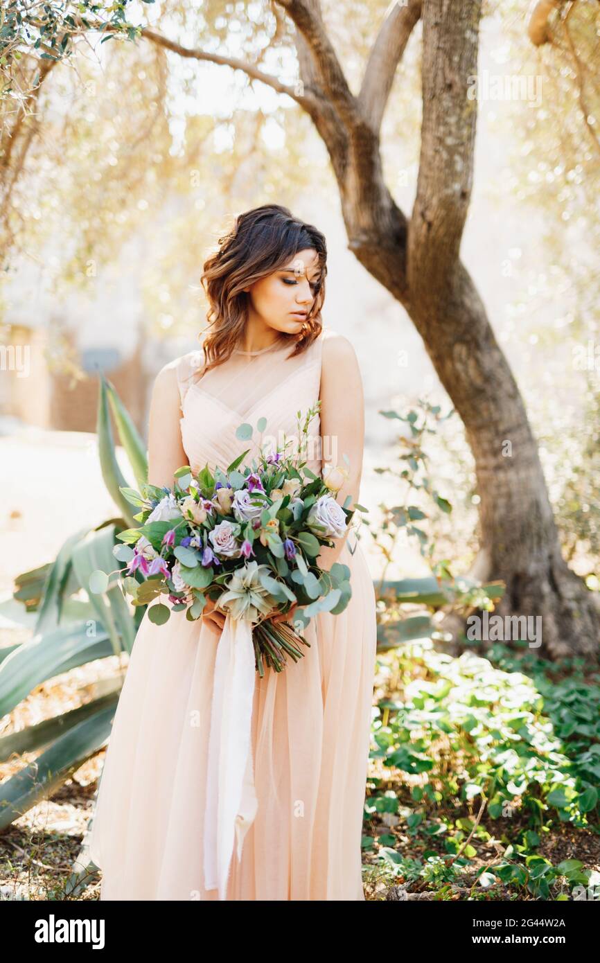 Cute bride in a beautiful dress stands pensively against a tree background with a gorgeous bouquet of flowers in her hands Stock Photo