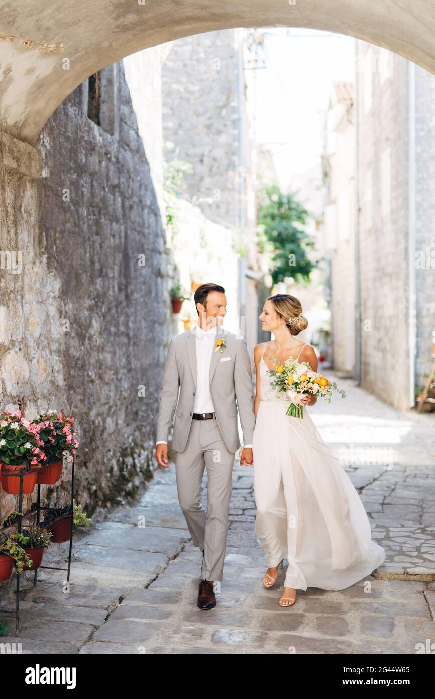 The bride and groom with a bouquet walk hand in hand along a cozy street of the old city under the arch and smile Stock Photo