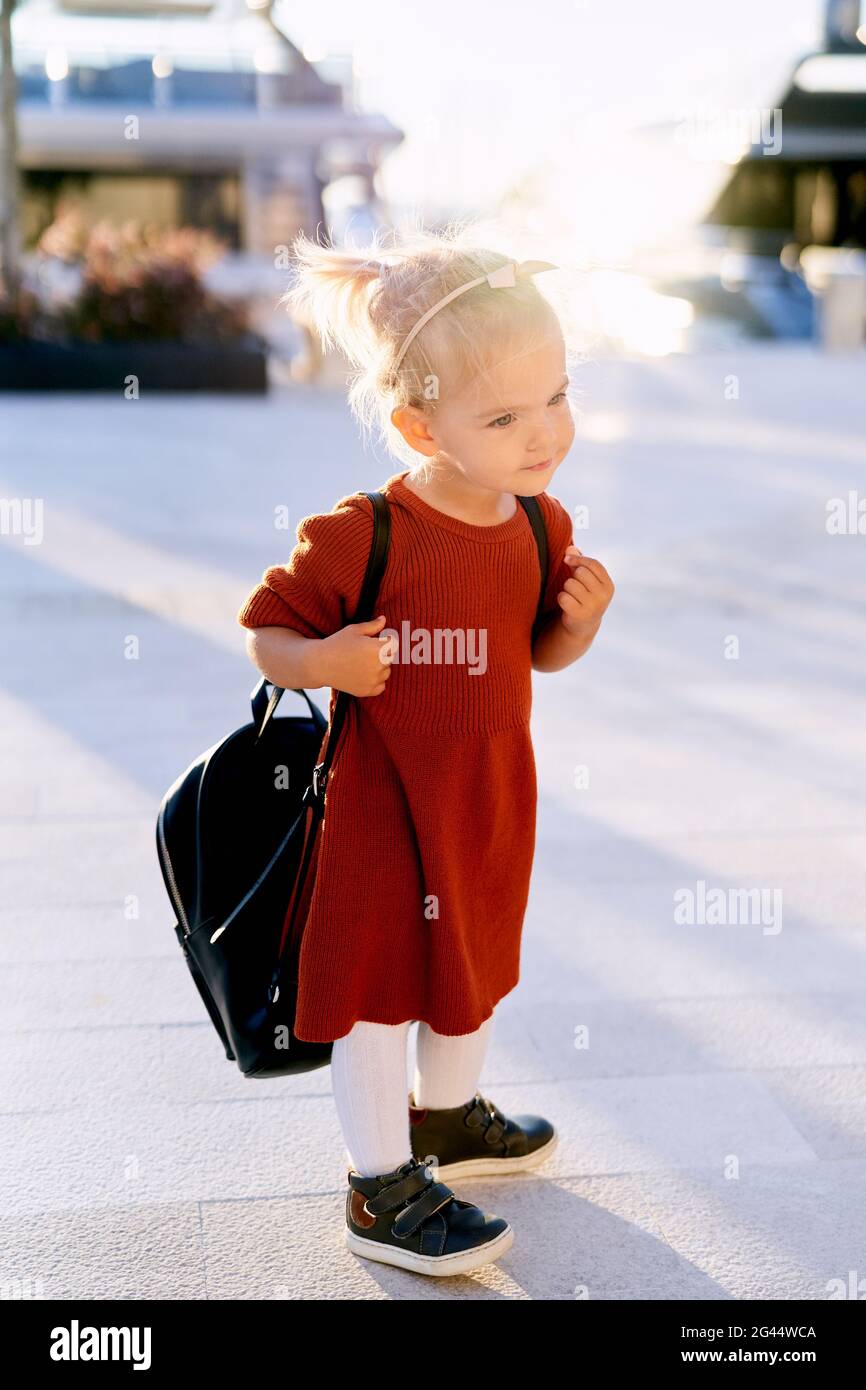Little girl in a terracotta dress is carrying a big black backpack on her back Stock Photo