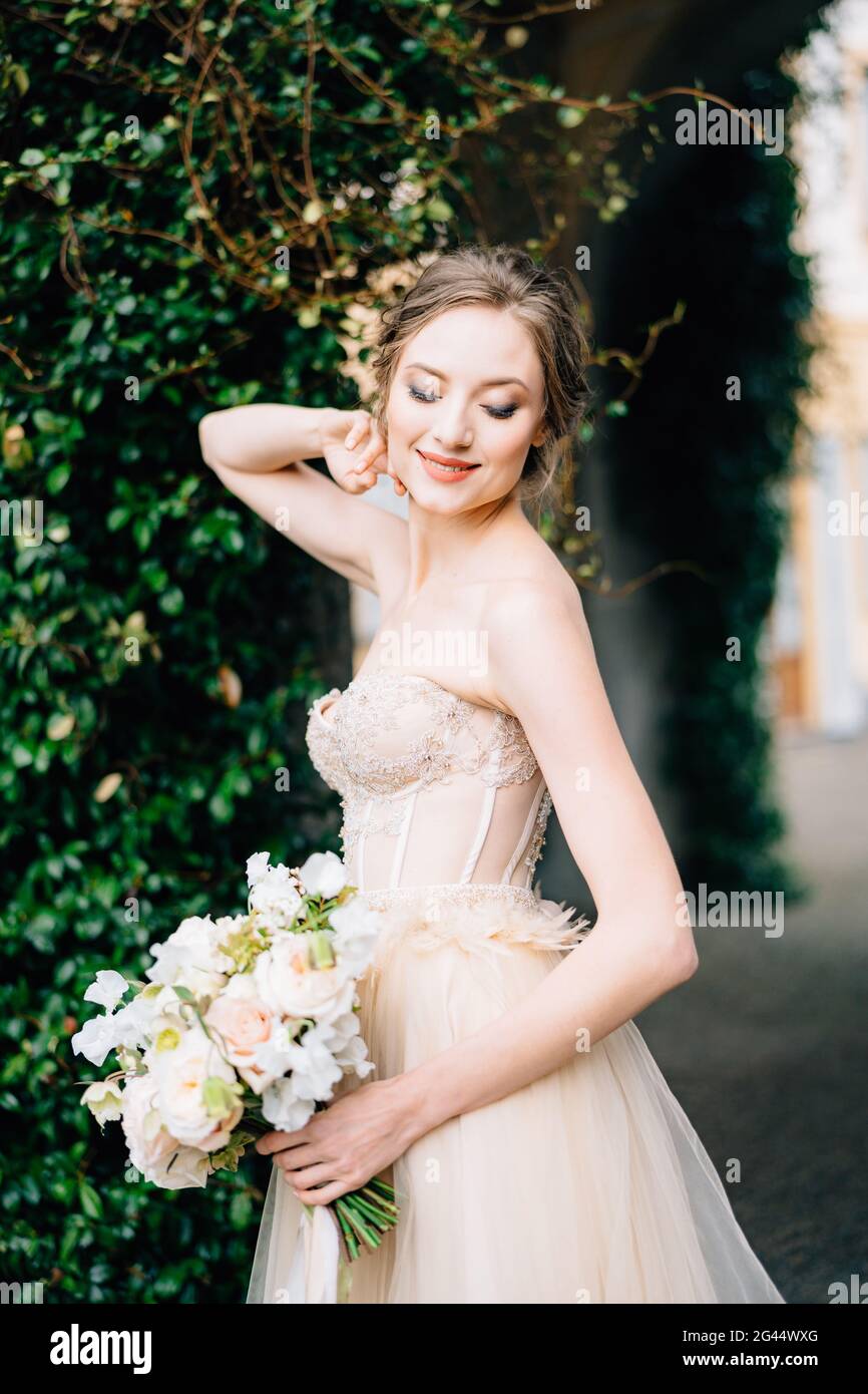 Smiling bride in an off-the-shoulder dress with a bouquet of pink flowers put her hand behind her head. Lake Como, Italy Stock Photo