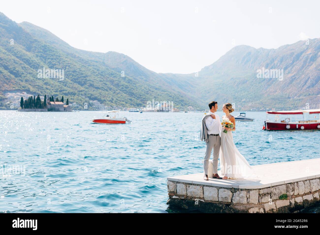 The bride and groom standing side by side on the pier in the Bay of Kotor, islands of Perast are behind them Stock Photo