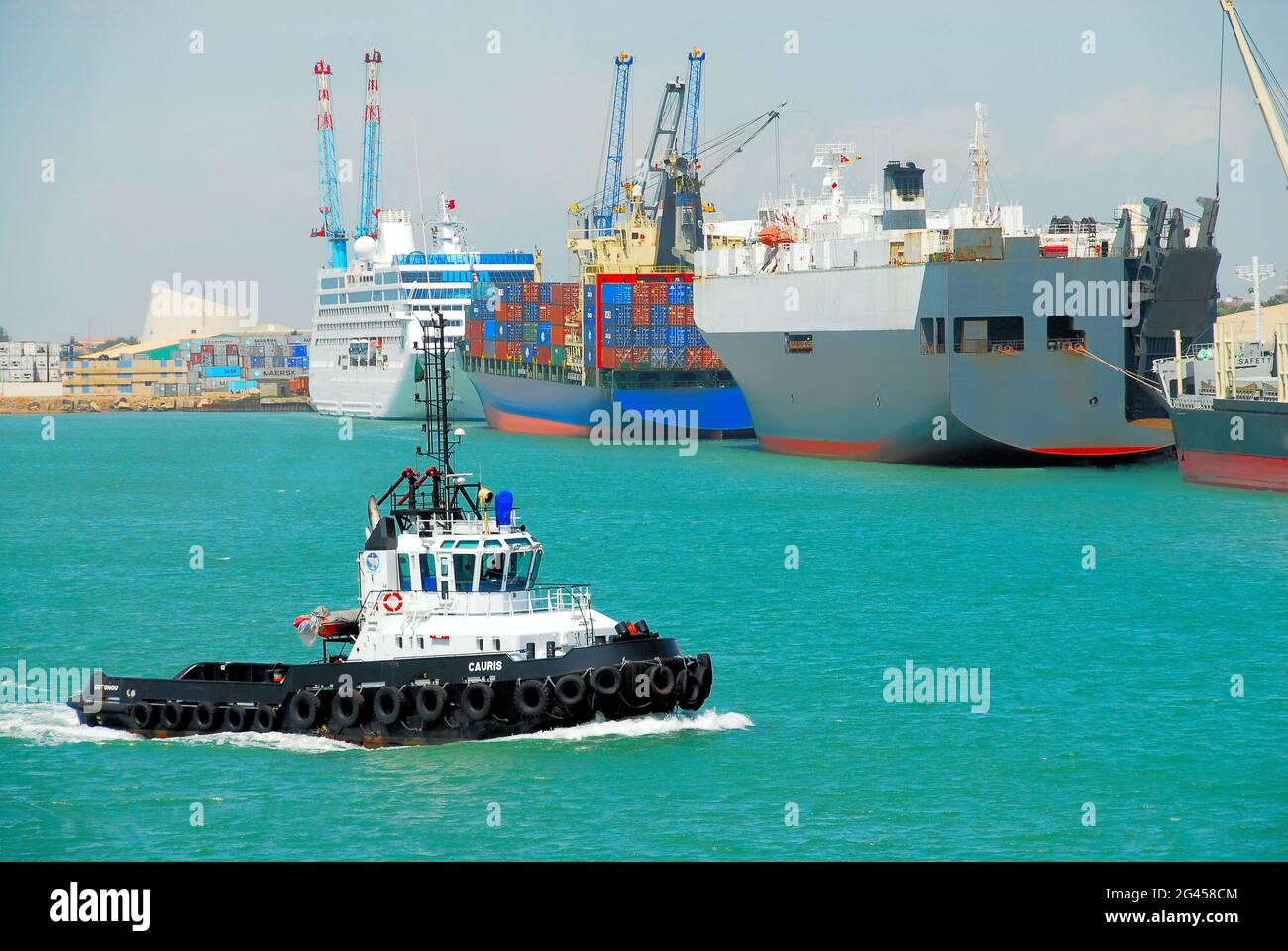 A diversity of ships docked in the turquoise waters at the Autonomous Port of Cotonou, Benin, West Africa while a tug boat travels in the foreground. Stock Photo