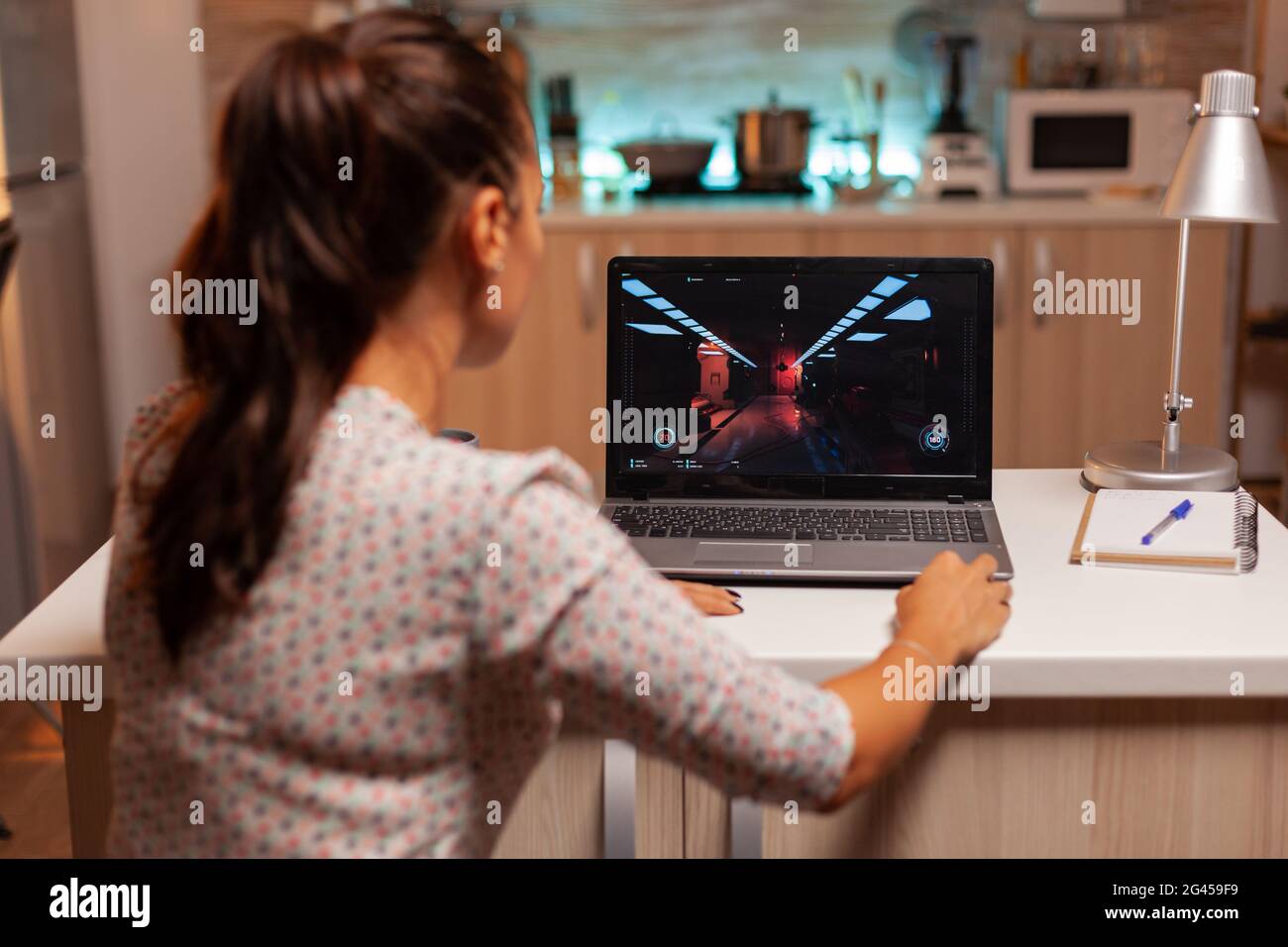 Lady playing games on laptop during night time during night time in home kitchen. Professional gamer playing online videop games on her personal computer. Geek cyber e-sport. Stock Photo