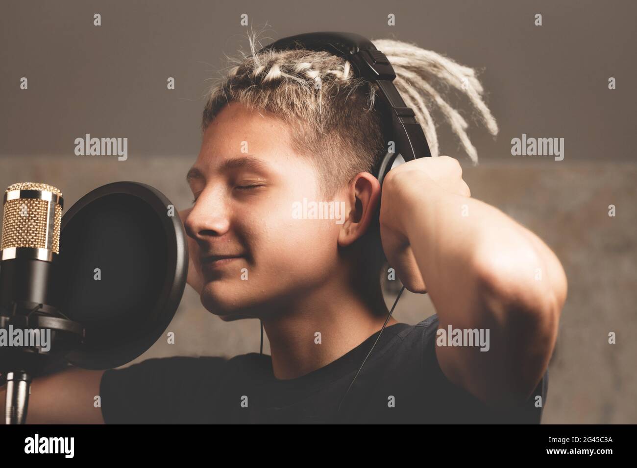Stylish positive guy with dreadlocks is recording a song in the studio. A young attractive singer in black studio headphones stands in front of a micr Stock Photo