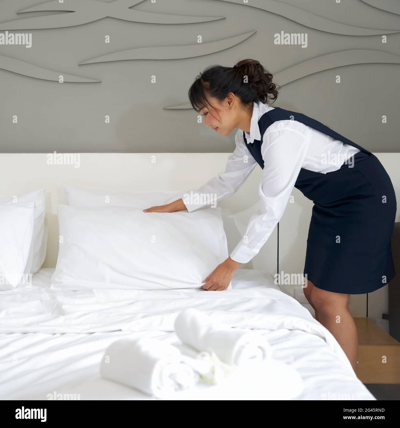 Young hotel maid making the bed. Hotel staff in blue uniform preparing room for guest Stock