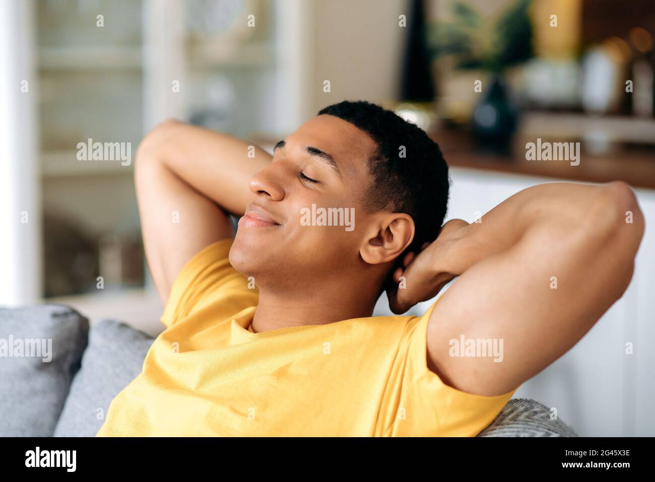 Relaxed satisfied calm hispanic guy in a yellow t-shirt, resting on the sofa at home with his eyes closed, throwing his hands behind his head, dreaming of a vacation, resting from work, smiling Stock Photo