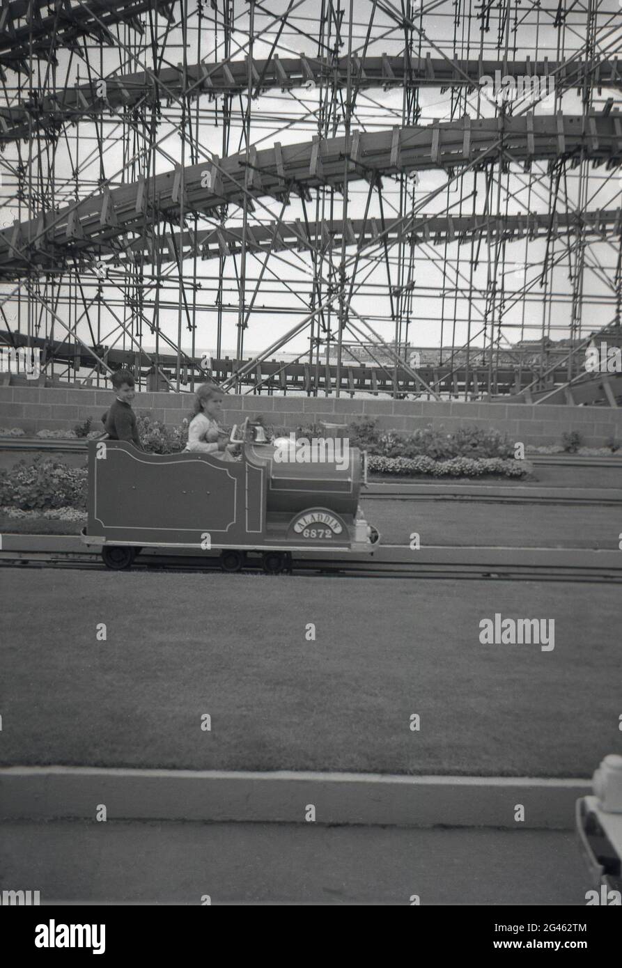 1962, historical two young children on a funfair train ride at Clacton-on-Sea, Essex, England, UK, with the big dipper of Clacton Pier in the background. Stock Photo