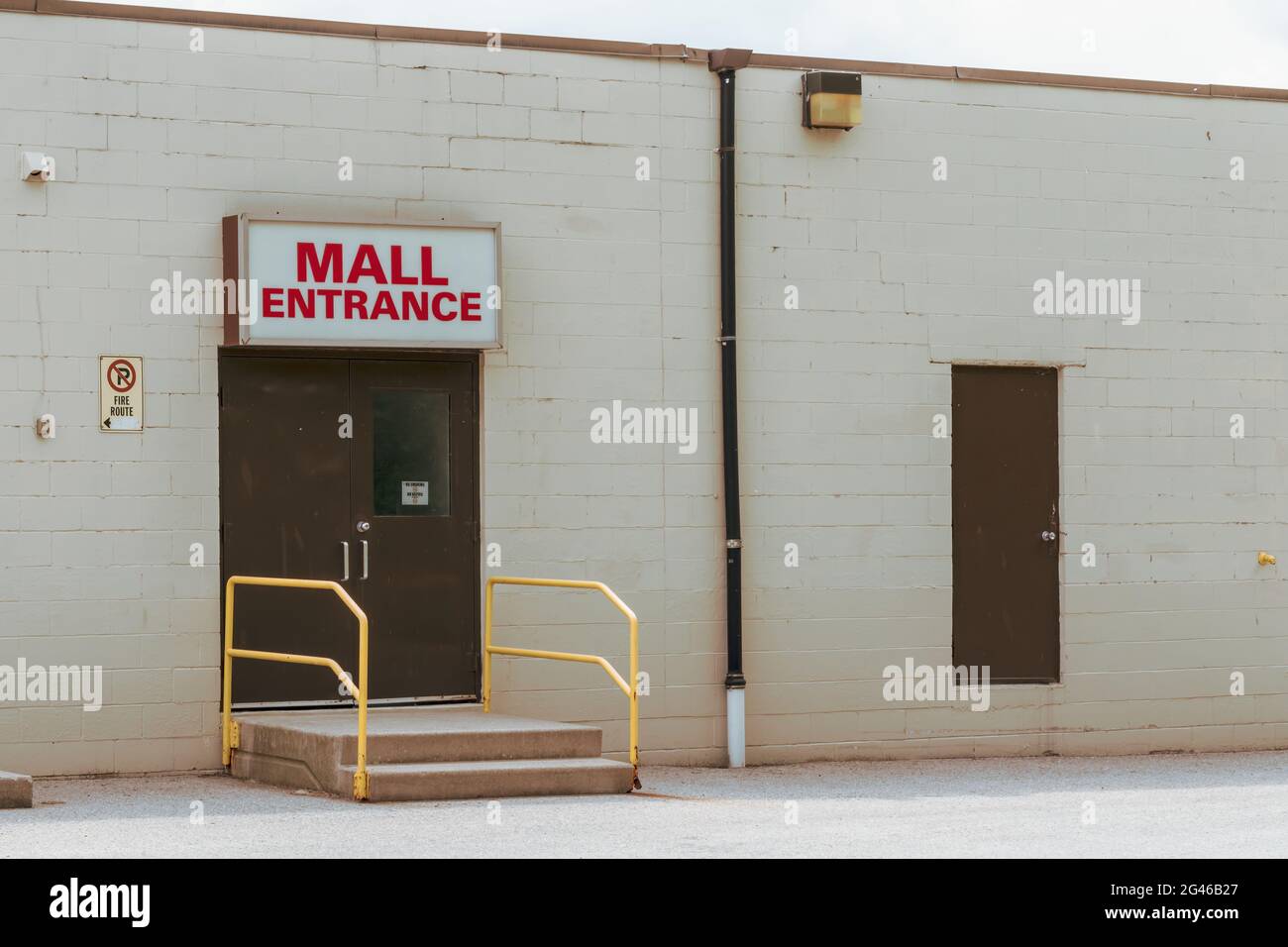 Ugly mall entrance - vintage filter. Brown doors. No smoking or vaping sign. Yellow handrails down two cement steps. White brick exterior wall. Stock Photo